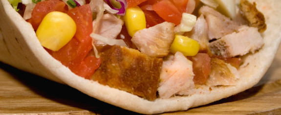 Close-up of a pita sandwich with grilled chicken, corn, tomatoes, and onions.
