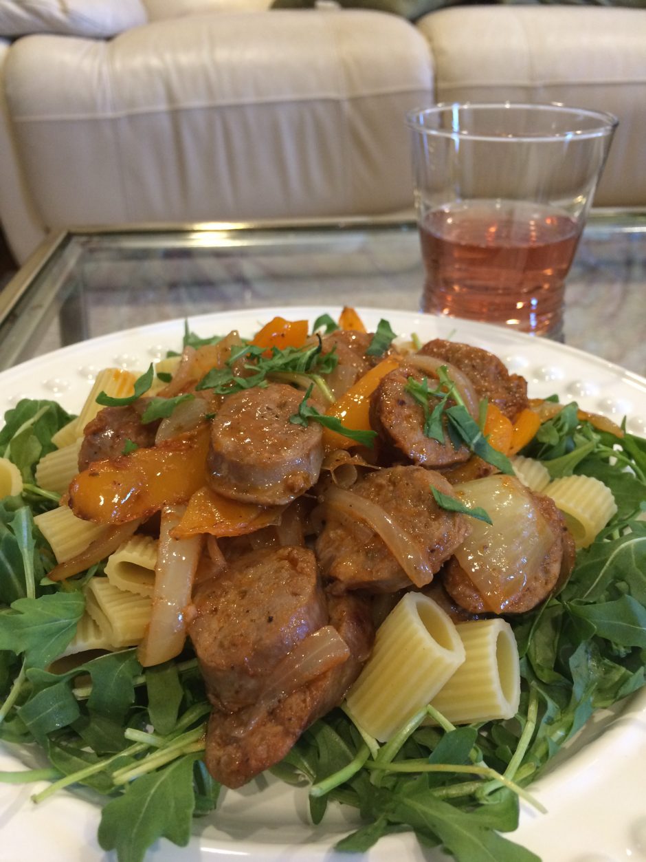 A plate of seasoned chicken with pasta on a bed of greens and a glass of beverage.