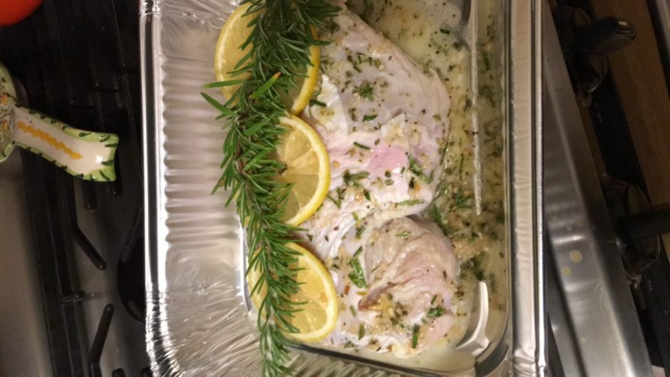 Baked fish garnished with lemon slices and rosemary in a foil tray.