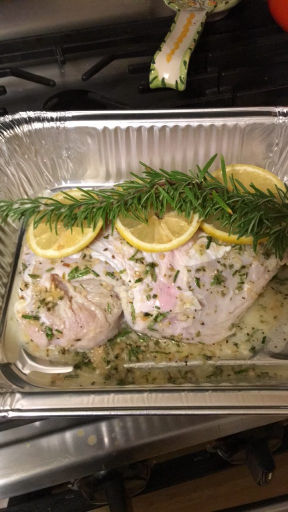 Baked fish garnished with lemon slices and rosemary in a foil tray.
